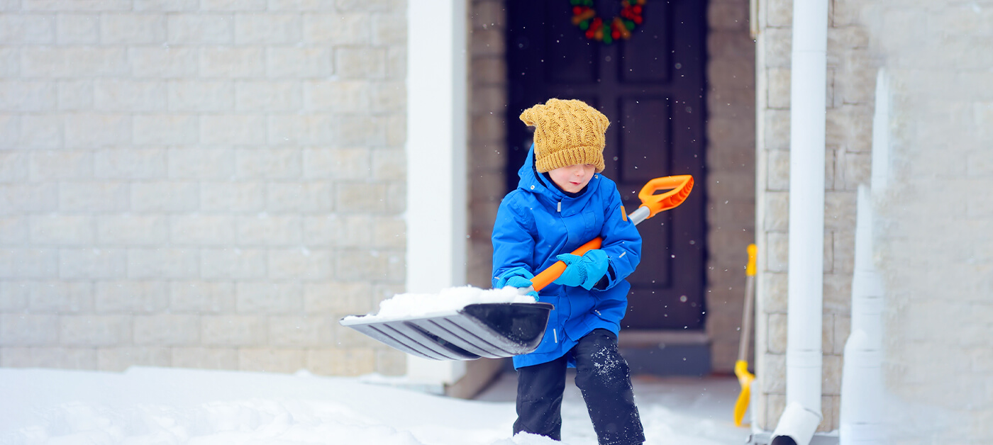 A young boy uses a large shovel to remove snow from the driveway in winter.