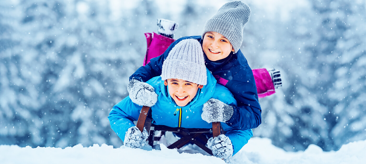 Two cheerful young boys are preparing to go sledding down a slope, face down, one on top of the other.