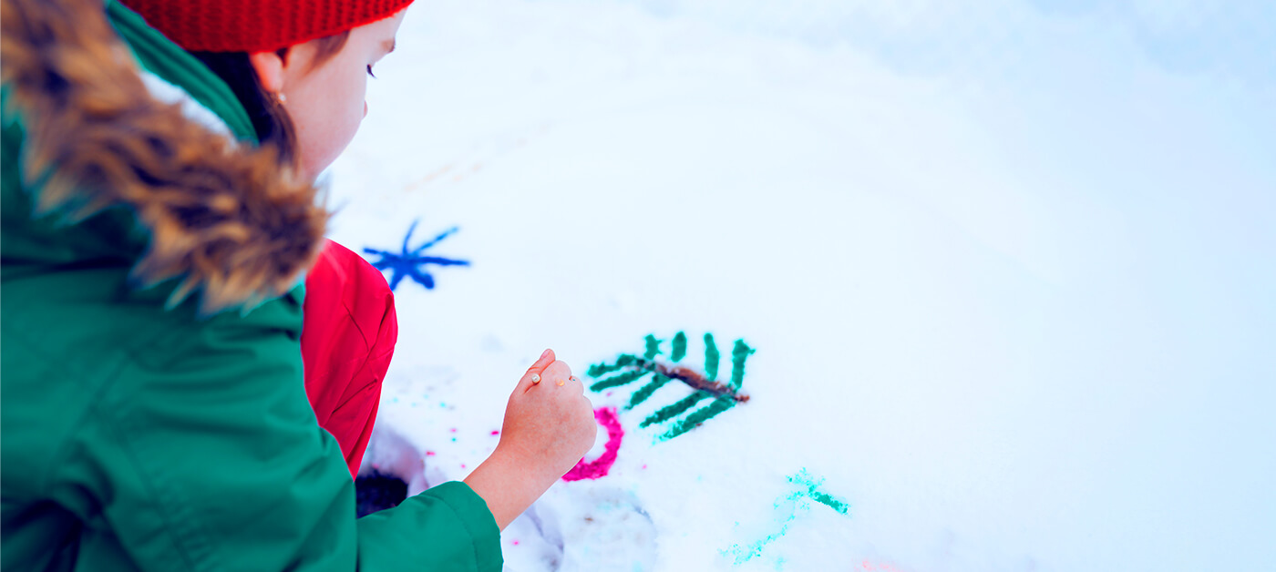 A little girl paints on the snow with a brush and washable gouache.