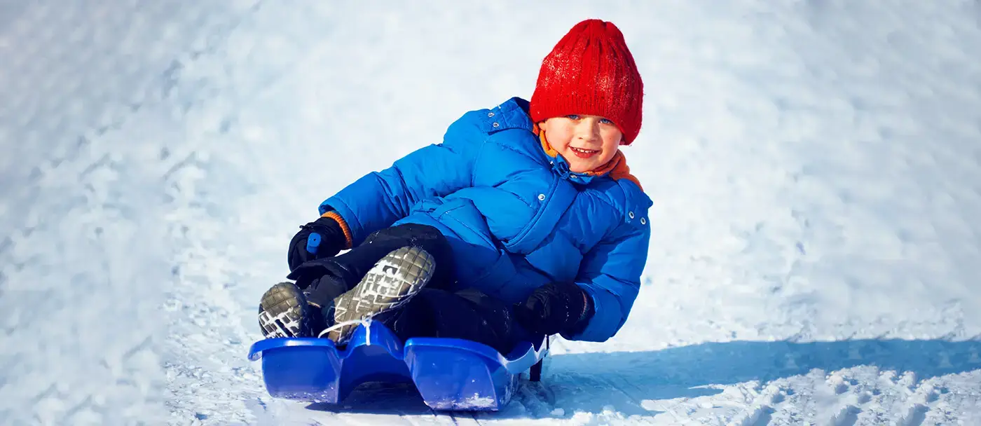 A young boy slides through the snow while seated in a sled.